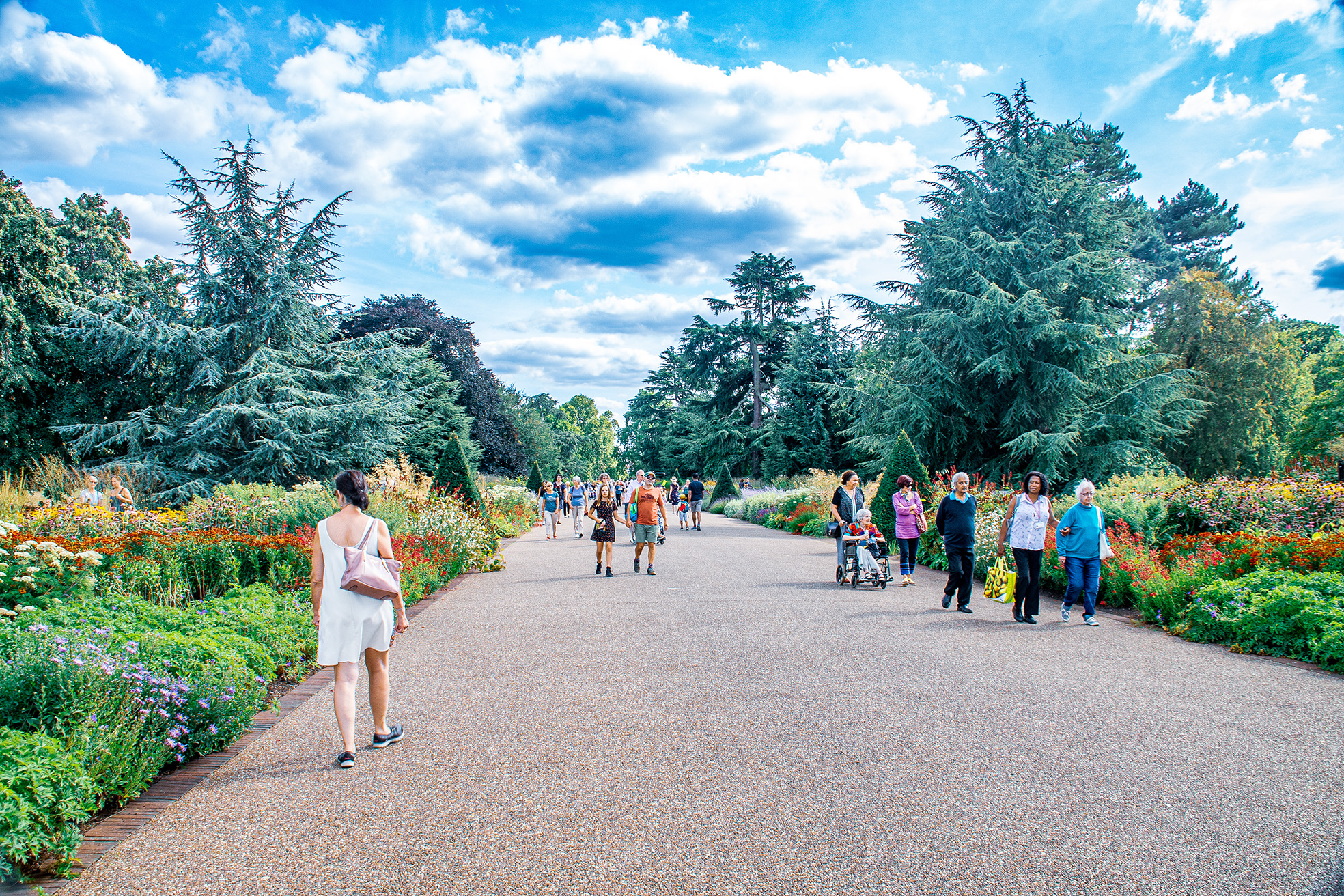 The image depicts a lively scene in a botanical garden with numerous people walking along a wide, gravel path. The path is lined with vibrant, colorful flowers and lush greenery on both sides, creating a picturesque and serene environment. Tall trees provide a canopy overhead, and the sky is bright with scattered clouds. People of various ages are seen enjoying the garden, some walking hand-in-hand, others pushing strollers, and a few carrying bags. The overall atmosphere is calm and inviting, suggesting a pleasant day spent outdoors.
