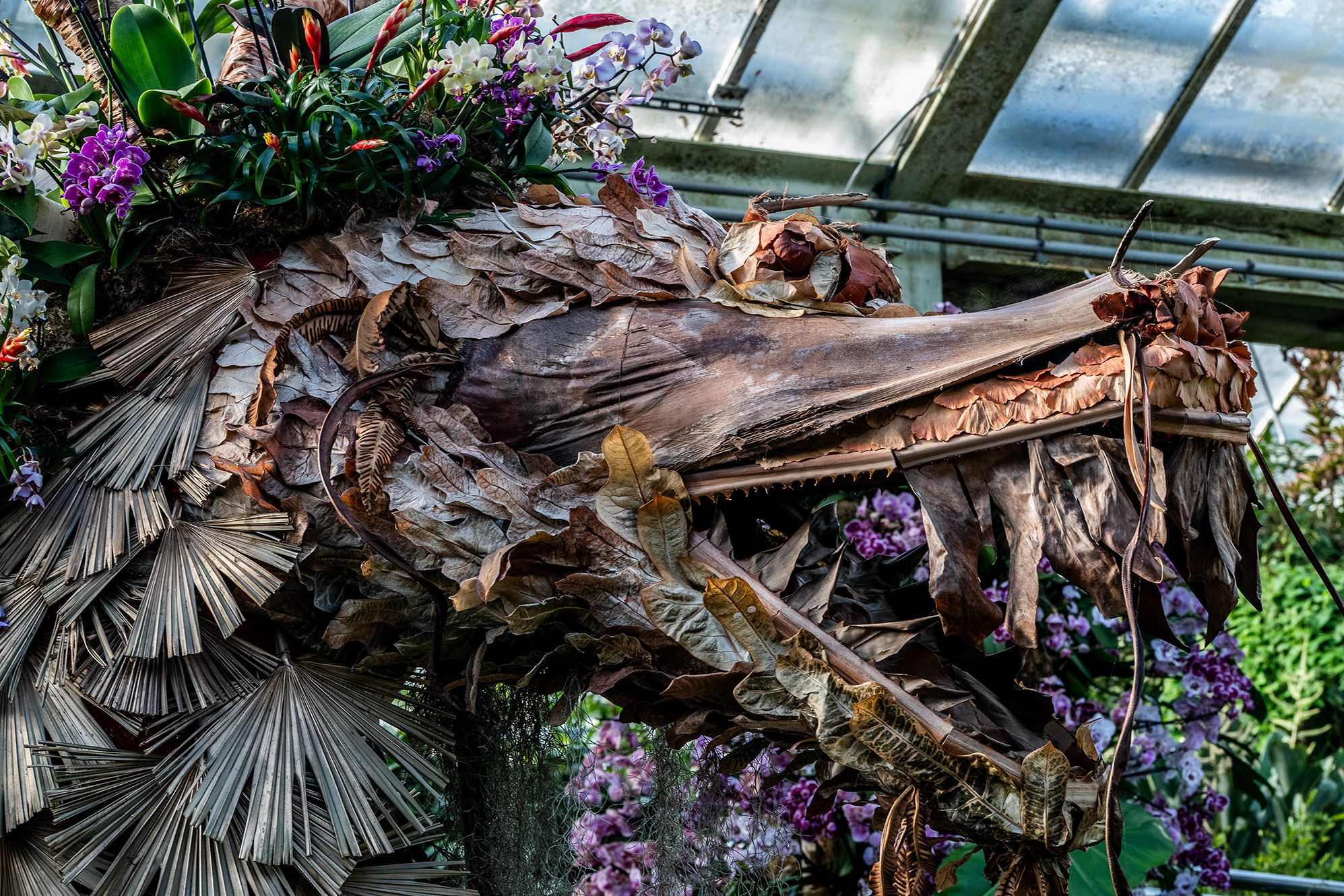The image depicts a close-up of a decorative structure made from dried leaves and branches, with various plants and flowers, including orchids and lilacs, growing on and around it. The structure appears to be part of a greenhouse or conservatory, as suggested by the metal framework and glass panels in the background.