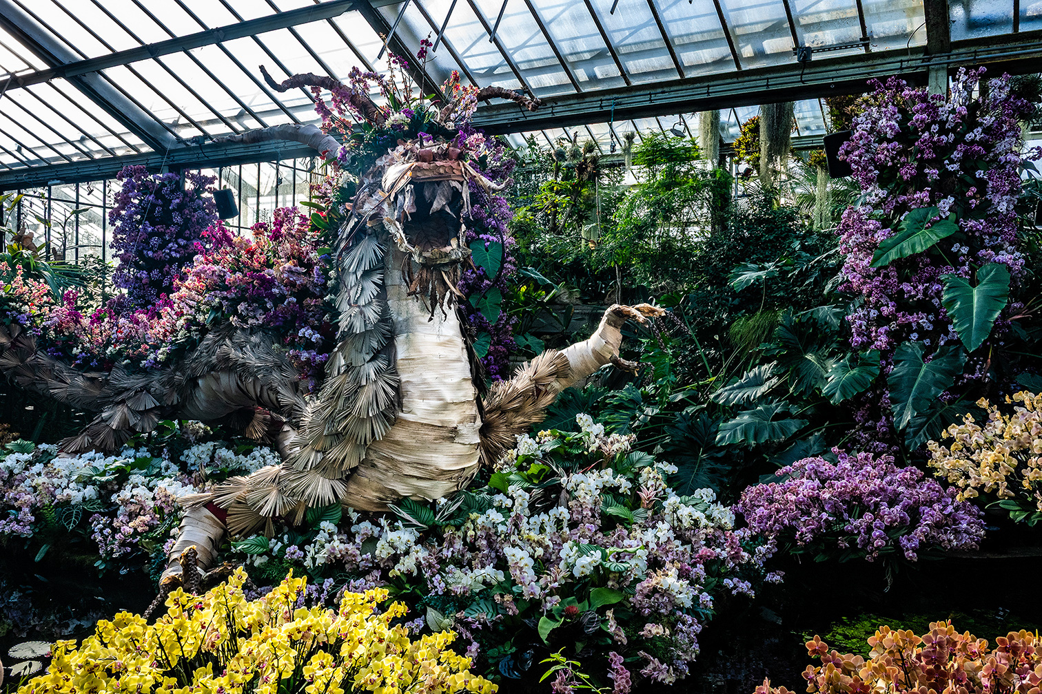 The image depicts a lush, indoor garden or greenhouse filled with a variety of vibrant flowers and plants. Central to the scene is a large, intricate sculpture made of natural materials like wood and leaves, resembling a mythical or abstract figure. The sculpture is surrounded by an array of colorful blooms, including yellow, purple, and white flowers, creating a vivid and lively atmosphere. The greenhouse structure is visible with its glass panels allowing natural light to illuminate the scene.