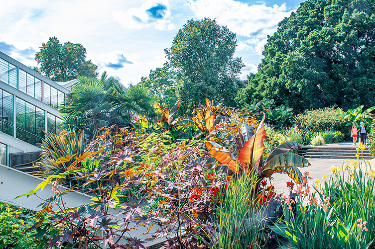 The image depicts a vibrant garden with a variety of colorful plants and flowers. There is a modern glass building on the left side, and a pathway leading through the garden with people walking in the background. The scene is set on a bright, sunny day with a clear blue sky.