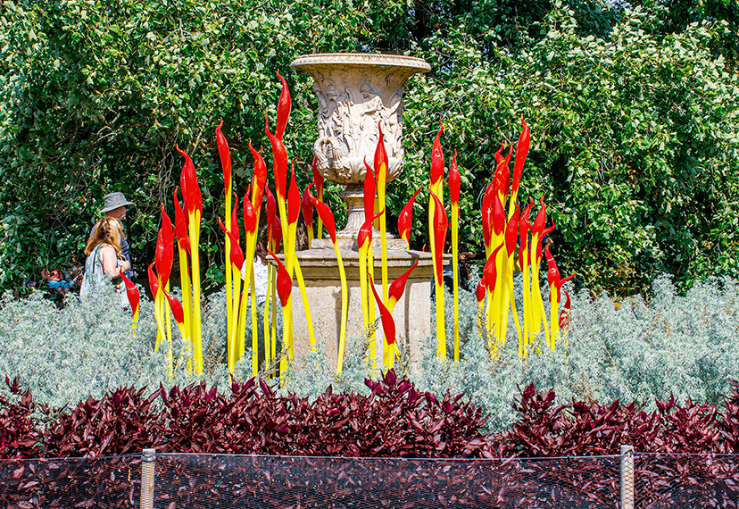 The image depicts an outdoor scene with a central stone urn surrounded by vibrant, flame-shaped sculptures in red and yellow. The sculptures are set within a well-maintained garden featuring various types of plants, including a bed of red and purple foliage in the foreground and a backdrop of lush green trees. A person wearing a hat and light-colored clothing is visible on the left side of the image, walking along a path.