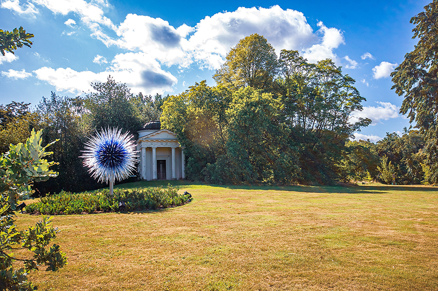 The image depicts a serene outdoor scene with a small, white, classical-style building situated in a lush garden. The building is surrounded by well-maintained greenery and trees under a clear blue sky with scattered clouds. In front of the building, there is a large, metallic, starburst sculpture adding a decorative element to the scene. The overall atmosphere is peaceful and inviting, suggesting a well-kept park or garden.
