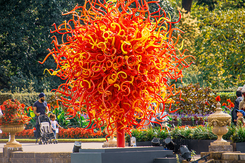 The image depicts a vibrant, colorful glass sculpture resembling a tree with red and orange tendrils, situated in an outdoor garden setting. People, including a person pushing a stroller, are seen walking around and admiring the sculpture amidst lush greenery and flowers.
