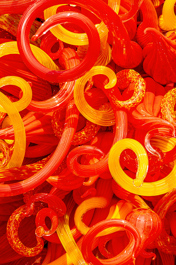The image displays a vibrant collection of red and orange glass pieces, including various shapes like loops, spirals, and spheres. The glass objects are arranged densely, creating a visually striking and colorful display.