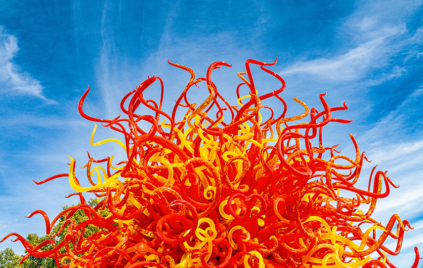 The document describes a vibrant, colorful sculpture set against a blue sky with some clouds. The sculpture is composed of numerous intertwined, flowing shapes in bright red, orange, and yellow colors, creating a dynamic and lively visual effect.