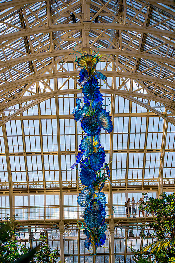 The image depicts a large, colorful glass sculpture hanging from the ceiling of a spacious, well-lit conservatory or greenhouse. The structure features a high, arched roof with a framework of wooden beams and numerous glass panes, allowing natural light to flood the interior. The glass sculpture is composed of numerous blue and green flower-like elements, creating a vibrant and eye-catching centerpiece. The conservatory is filled with various plants and greenery, and a few people can be seen observing the sculpture and the surroundings.