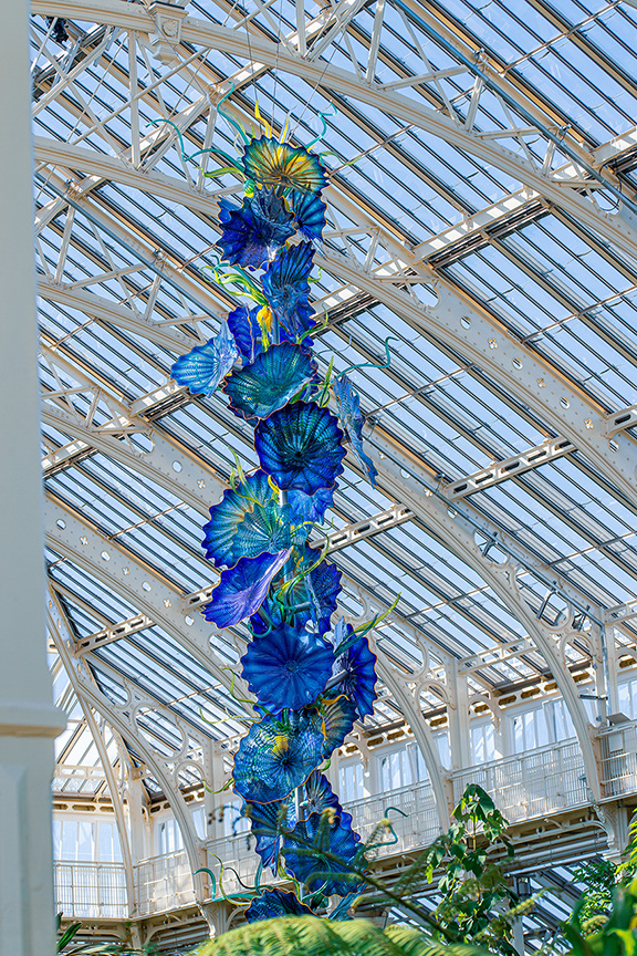 The image showcases a vibrant, large glass sculpture hanging from the ceiling of a greenhouse or conservatory. The sculpture is composed of blue and green glass elements resembling flowers and leaves. The structure of the building features a grid of glass panels allowing natural light to illuminate the interior. The surrounding area includes various plants and balconies, suggesting a multi-level environment.
