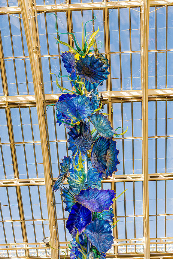 The image depicts a vibrant, colorful glass sculpture resembling a flower arrangement, hanging from a metal grid structure. The sculpture features a variety of blue, green, and yellow hues, with petals and leaves intricately designed. The background shows a clear blue sky, suggesting the sculpture is located in an outdoor or partially enclosed space with a transparent roof.