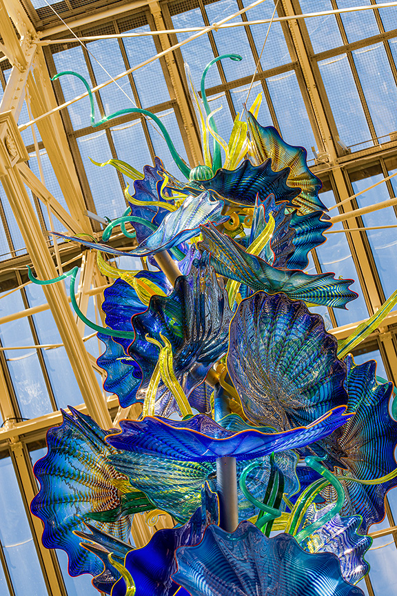 The image depicts a vibrant and intricate glass sculpture, likely a chandelier, with a variety of blue, green, and yellow hues. The sculpture features numerous flower-like forms and is suspended within a large, glass-roofed structure, allowing natural light to illuminate its vivid colors and detailed craftsmanship.