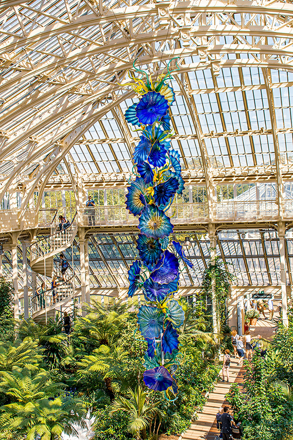 The image depicts a large, colorful glass sculpture suspended in a greenhouse-like structure. The sculpture features vibrant blue and green flower-like forms arranged in a vertical pattern. The greenhouse has a high, arched ceiling made of glass and metal, allowing natural light to illuminate the space. Lush greenery and various plants surround the sculpture, creating a vibrant and lively atmosphere. People can be seen walking and observing the surroundings on different levels of the structure.