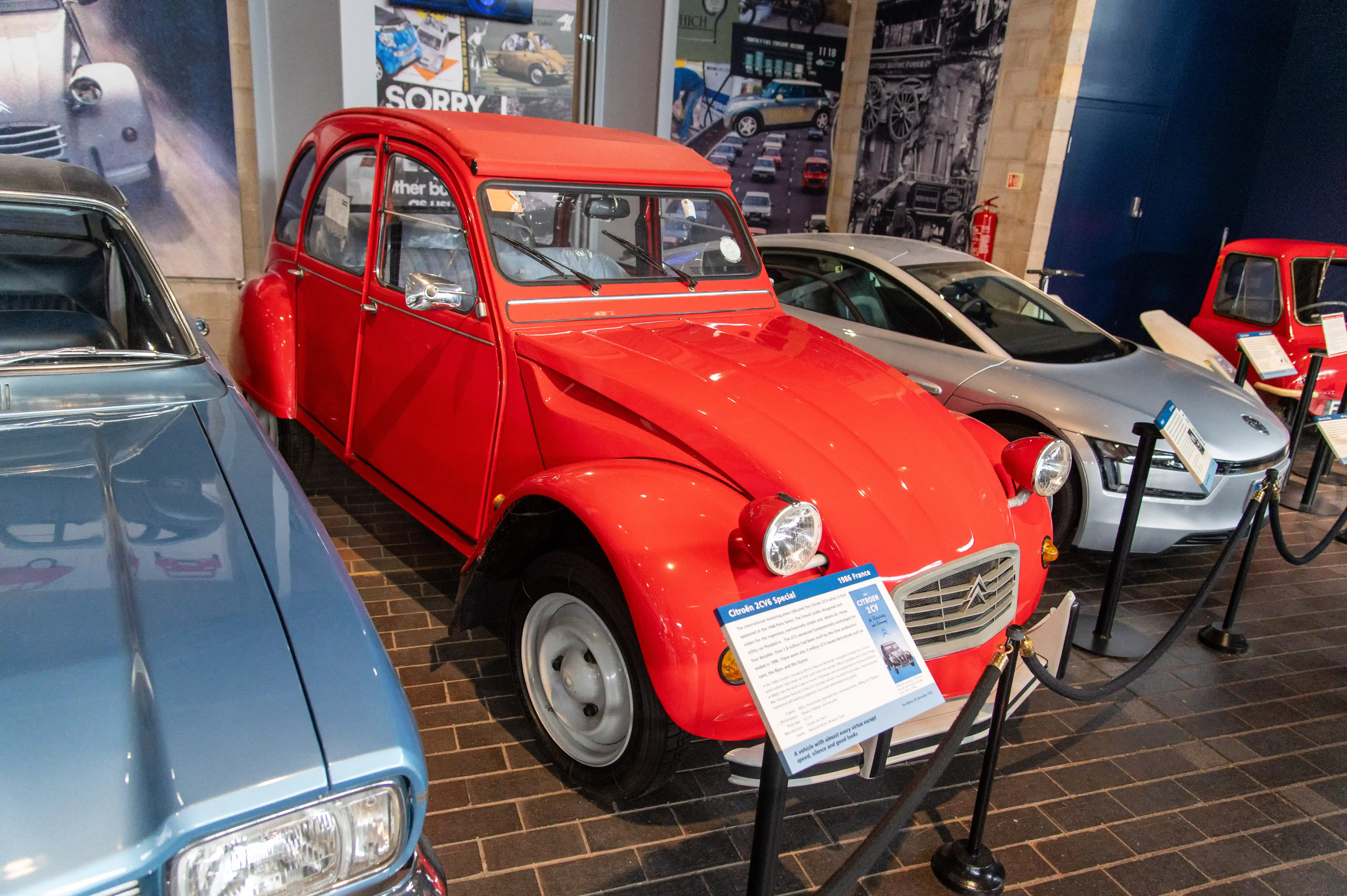 The image shows a display of classic Citroën cars, prominently featuring a red 1966 Citroën 2CV Special in the center. The cars are exhibited indoors, with informational placards in front of them, suggesting they are part of a museum or an exhibition. The setting includes various vintage car-related posters and photographs on the walls, enhancing the historical context of the display.