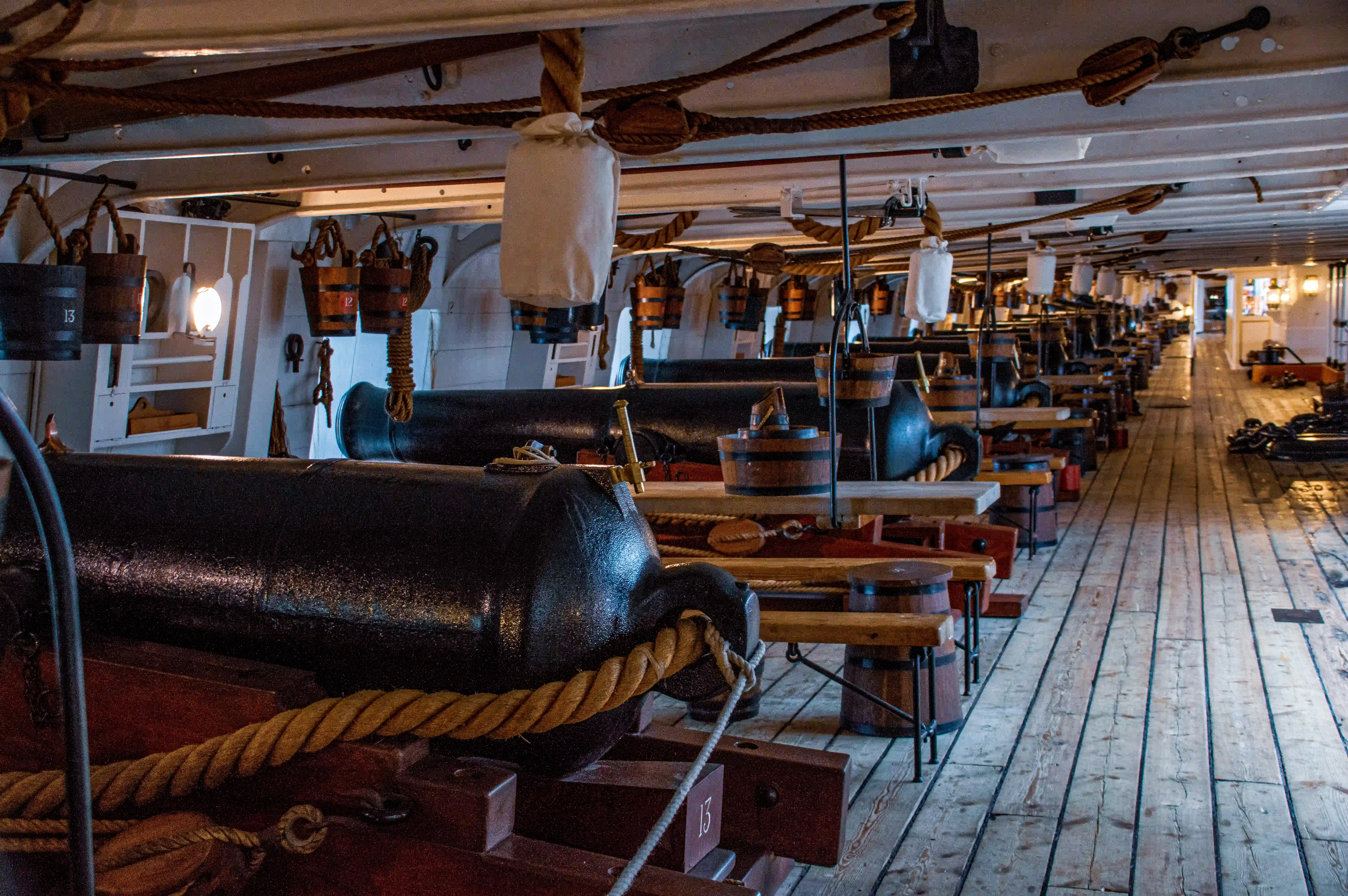The image depicts the interior of an old ship, specifically the gun deck. It shows a row of cannons lined up along the deck, each secured with ropes and positioned near wooden benches and various equipment. The deck is made of wooden planks, and the ship's structure includes white walls and beams. The lighting is dim, with some light coming from small windows and lanterns. The overall scene gives a sense of historical naval architecture and the operational environment of a warship.