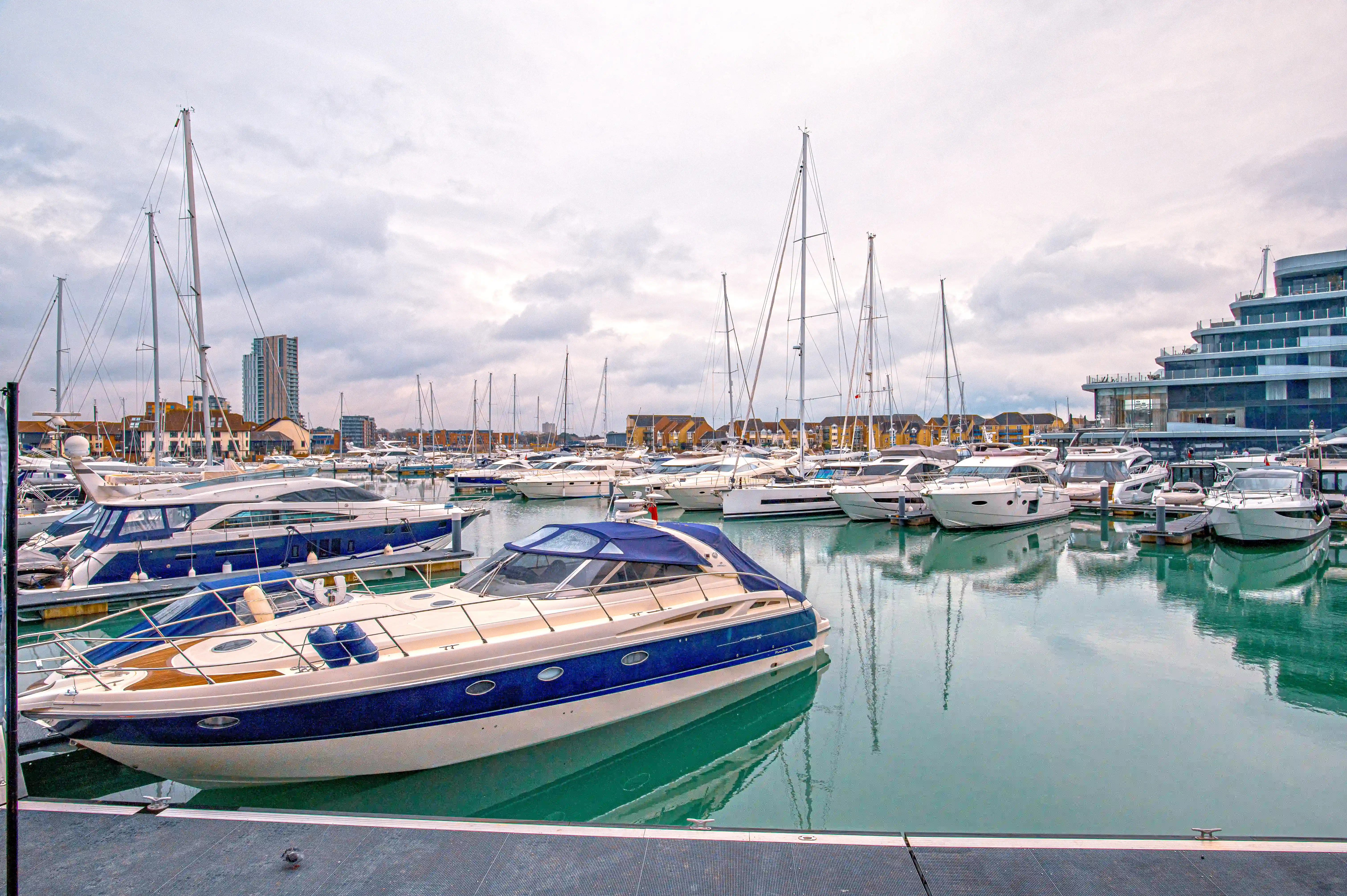 The image depicts a modern marina filled with various types of boats and yachts docked in calm, greenish-blue water. The marina is surrounded by contemporary buildings and residential structures, indicating a well-developed waterfront area. The sky is overcast, suggesting a cloudy day. The overall scene conveys a sense of tranquility and modernity.