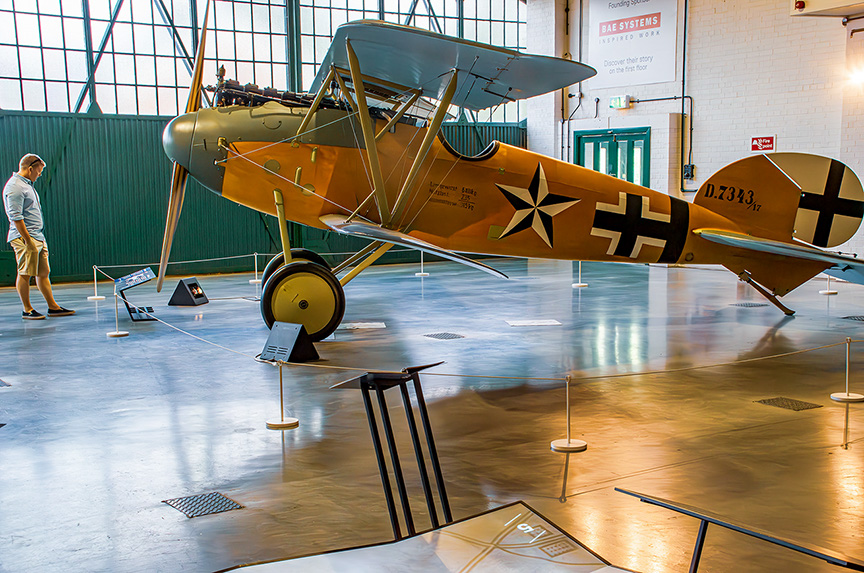 The image shows a vintage military aircraft displayed in a museum setting. The aircraft is painted in a yellow and green camouflage scheme with black crosses on its wings and fuselage, indicative of World War II-era German Luftwaffe aircraft. A man is standing nearby, observing the aircraft. The plane is cordoned off with barriers and informational placards, suggesting it is part of an exhibit. The setting appears to be a large, well-lit hangar with a high ceiling and large windows