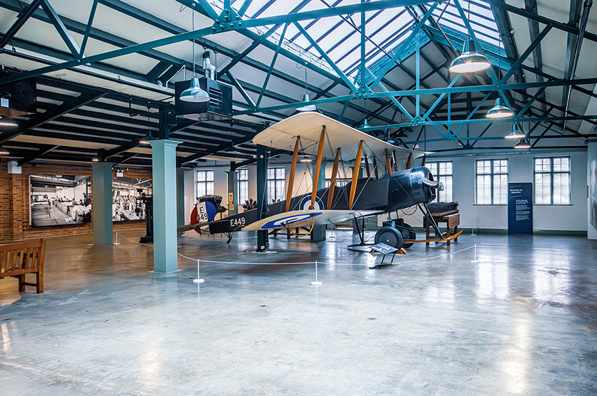 The image depicts an aviation museum exhibit featuring a vintage biplane. The museum has a high, industrial-style ceiling with exposed beams and large windows, allowing natural light to illuminate the space. The biplane is prominently displayed in the center, with informational displays nearby. The floor is polished concrete, and there are barriers around the exhibit to prevent visitors from touching the aircraft. The overall atmosphere is one of historical preservation and educational display.