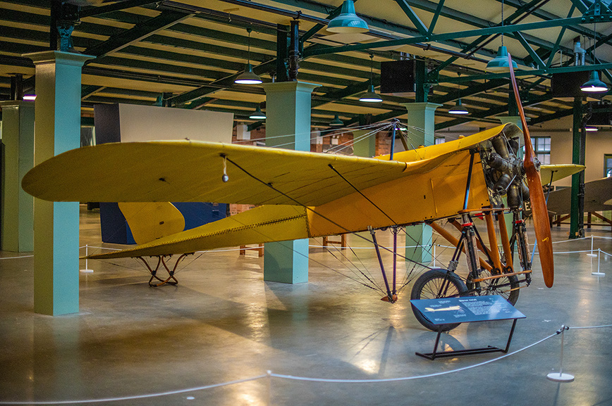 The image shows a vintage yellow biplane on display in a museum. The aircraft is mounted on a stand with a sign in front of it, providing information about the plane. The biplane features a wooden propeller and is suspended in a manner that allows viewers to see it from multiple angles. The museum setting includes high ceilings, green columns, and ample lighting, creating a spacious and well-lit environment for the exhibit.