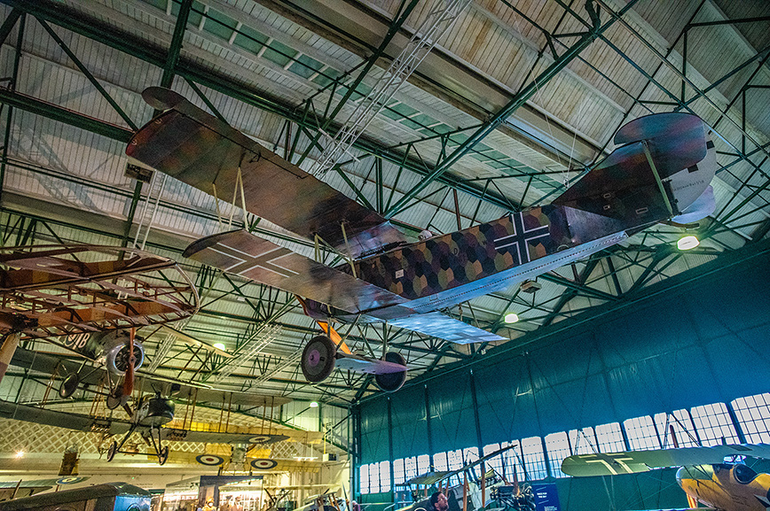 The image depicts a museum showcasing historical aircraft. The central focus is on a biplane with a distinctive iron cross marking on its side, indicative of German World War I-era aircraft. The plane is suspended from the ceiling, highlighting its design and markings. Other aircraft and aviation artifacts are visible in the background, suggesting a well-curated collection of historical significance.