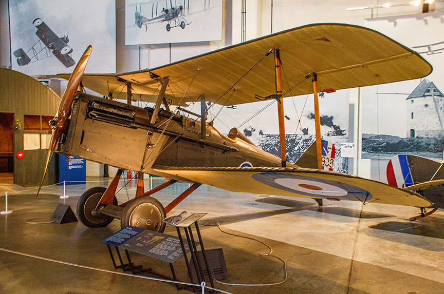 The image shows a vintage biplane on display in a museum. The aircraft is a Sopwith Camel, a type of single-seat biplane fighter flown by the British during World War I. The plane is painted in a brown camouflage scheme with a roundel insignia on the side of the fuselage and the underside of the wings. The display includes informational placards providing details about the aircraft's history and specifications. The background features historical photographs and a diorama of a World War I battlefield.