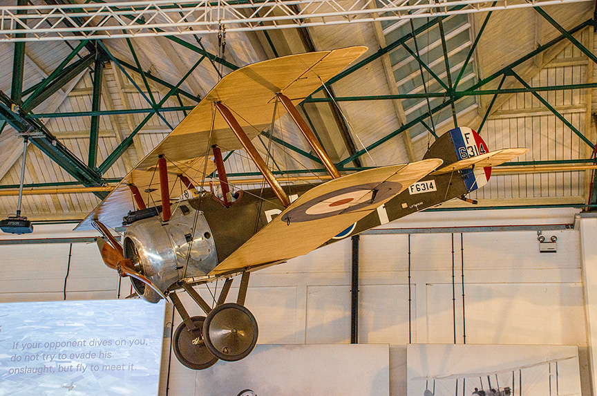 The image shows a vintage biplane, specifically a Sopwith Camel, suspended from the ceiling of a museum or exhibition hall. The aircraft is painted in a camouflage scheme typical of World War I era planes, featuring a roundel on the side and various markings. The biplane has a distinctive twin-wing structure with a fabric-covered frame. The setting appears to be an indoor aviation museum, with informational displays visible in the background.