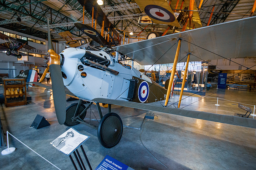 The image depicts a vintage biplane on display in a museum. The aircraft is painted in a light blue color scheme with distinctive roundels on its wings and fuselage. The biplane features a single engine with a large propeller at the front, and it is supported by a landing gear with large wheels. The museum setting includes other aircraft and informational displays in the background, indicating a focus on aviation history.