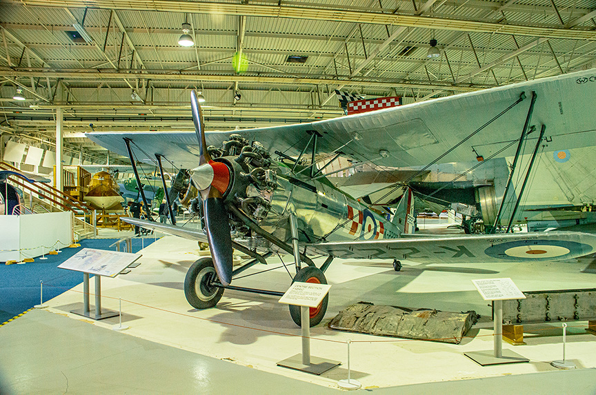 The image depicts an aircraft displayed in a museum setting. The aircraft is a vintage biplane with a radial engine, featuring distinctive roundels on its wings. It is positioned on a concrete floor with informational placards placed nearby, providing details about the aircraft. The museum space is spacious with high ceilings and industrial lighting, and other aircraft and exhibits are visible in the background.