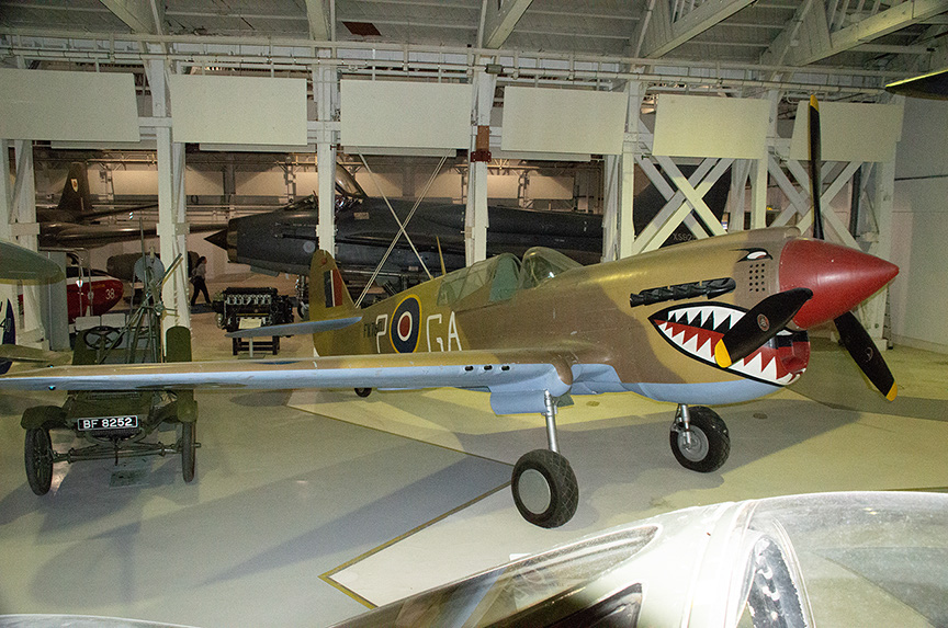 The image shows a vintage military aircraft displayed in a museum. The aircraft features a distinctive shark mouth design on its nose, characteristic of World War II fighter planes. The plane is positioned on a concrete floor with other aircraft visible in the background. The setting appears to be an indoor hangar with high ceilings and industrial lighting.