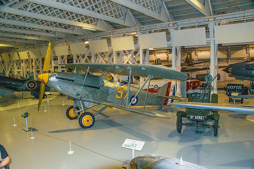 The image depicts an aircraft displayed in a museum. The aircraft is a vintage biplane with a yellow propeller and is marked with various insignia and numbers. It is situated in a large, well-lit hangar with other aircraft and exhibits visible in the background. The plane is supported by stands and has informational placards nearby, indicating it is part of a historical exhibit.