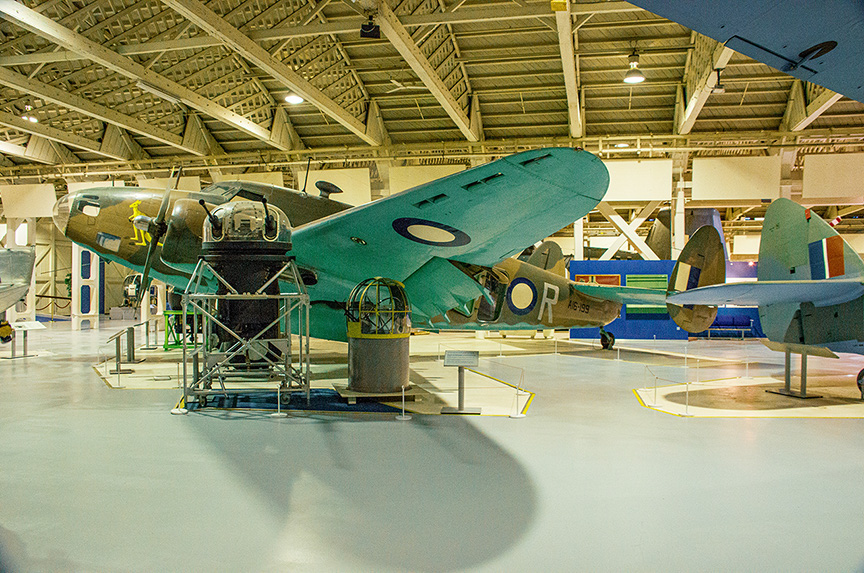 The image depicts an aircraft museum with several vintage planes on display. The central aircraft is painted in a distinctive blue-green color with roundels on its wings and fuselage. The planes are positioned on the floor with barriers around them, indicating they are part of an exhibit. The museum has a high ceiling with exposed beams and ample lighting, creating a well-lit environment for viewing the aircraft.