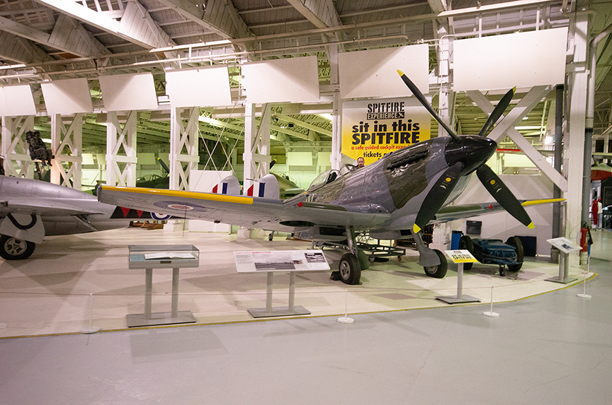 The image depicts an indoor aviation museum exhibit featuring a Spitfire aircraft. The museum has a high ceiling with exposed beams and houses several aircraft on display. The Spitfire is prominently positioned with informational placards in front of it, and there is a sign encouraging visitors to sit in the Spitfire. The environment is well-lit, and the museum appears to be spacious and organized.