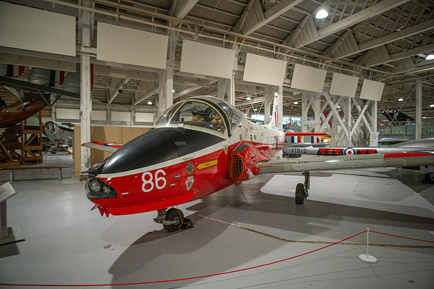 The image shows a red and white aircraft with the number 86 on its nose, displayed in a museum-like setting. The aircraft is positioned on a smooth floor with a red rope barrier in front of it, indicating it is part of an exhibit. The interior of the facility is spacious with high ceilings and large windows, allowing natural light to illuminate the aircraft. Other aircraft and exhibits are visible in the background, suggesting a diverse collection of aviation artifacts. The overall atmosphere is one of preservation and display, likely intended for educational or historical purposes.