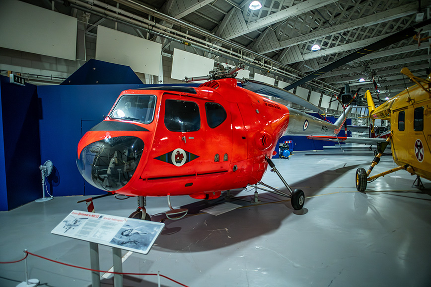 The image shows a red helicopter displayed in a museum setting. The helicopter is positioned on a platform with an informational placard in front of it. The museum is spacious with high ceilings and other aircraft visible in the background. The helicopter has a distinctive design with a bubble-like front and a black and white insignia on its side. The area is cordoned off with a red rope barrier.