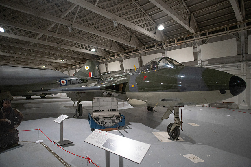 The image shows a vintage military jet aircraft displayed in a museum. The aircraft is positioned in a large, well-lit hangar with a high ceiling. Various informational placards are placed around the aircraft, providing details about it. There is a person sitting on a bench nearby, and the aircraft is equipped with a missile or rocket under its fuselage. The overall setting suggests an educational exhibit focused on aviation history.