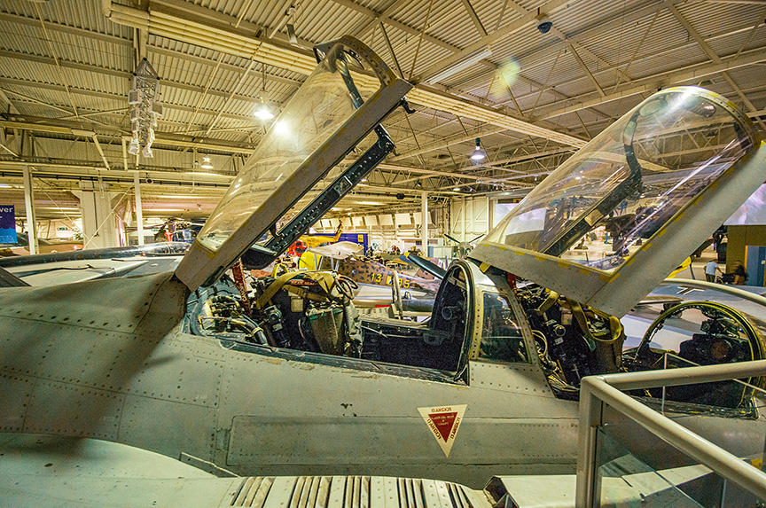 The image shows the cockpit of a military aircraft on display in a museum. The cockpit is open, revealing various controls and instruments. The aircraft is housed in a large hangar with high ceilings and other exhibits visible in the background. The cockpit canopy is open, providing a clear view of the interior. The aircraft appears to be well-preserved and is part of an aviation exhibit.