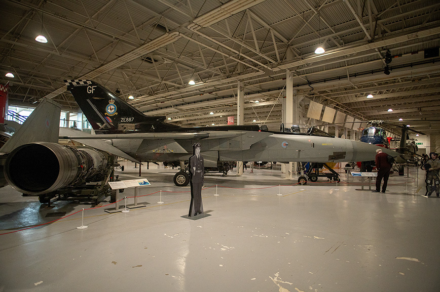 The image depicts an aircraft exhibit in a large hangar. The main focus is on a military jet with the tail markings 'GF' and 'ZE387'. There is a statue of a person in a military uniform standing in front of the jet. The hangar contains other aircraft and informational displays, with a few people observing the exhibits.