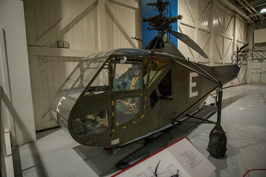 The image shows a military helicopter on display in a museum or exhibition setting. The helicopter is painted in a camouflage pattern and has a distinctive design with a bubble-like cockpit. It is mounted on a stand with an informational placard in front of it, providing details about the aircraft. The setting appears to be an indoor hangar with high ceilings and other aircraft or exhibits visible in the background.
