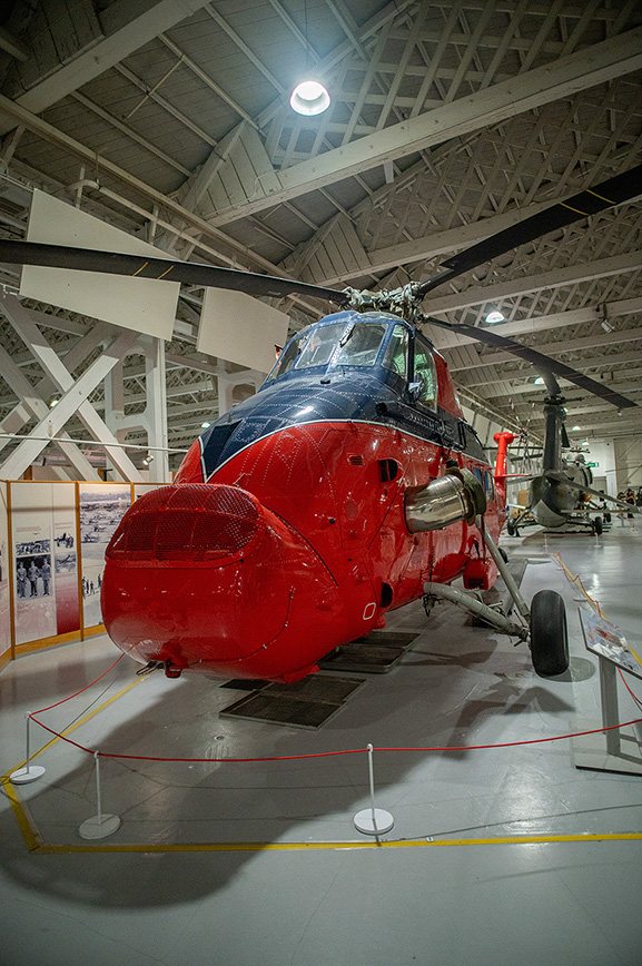The image depicts a red helicopter on display in a museum-like setting. The helicopter is cordoned off with red ropes and stanchions, indicating it is part of an exhibit. The environment is spacious with a high ceiling and industrial lighting, suggesting it is housed in a large hangar or exhibition hall. There are informational displays and other aircraft visible in the background, further emphasizing the museum context. The helicopter appears to be well-maintained and polished, showcasing its vibrant red color and sleek design.