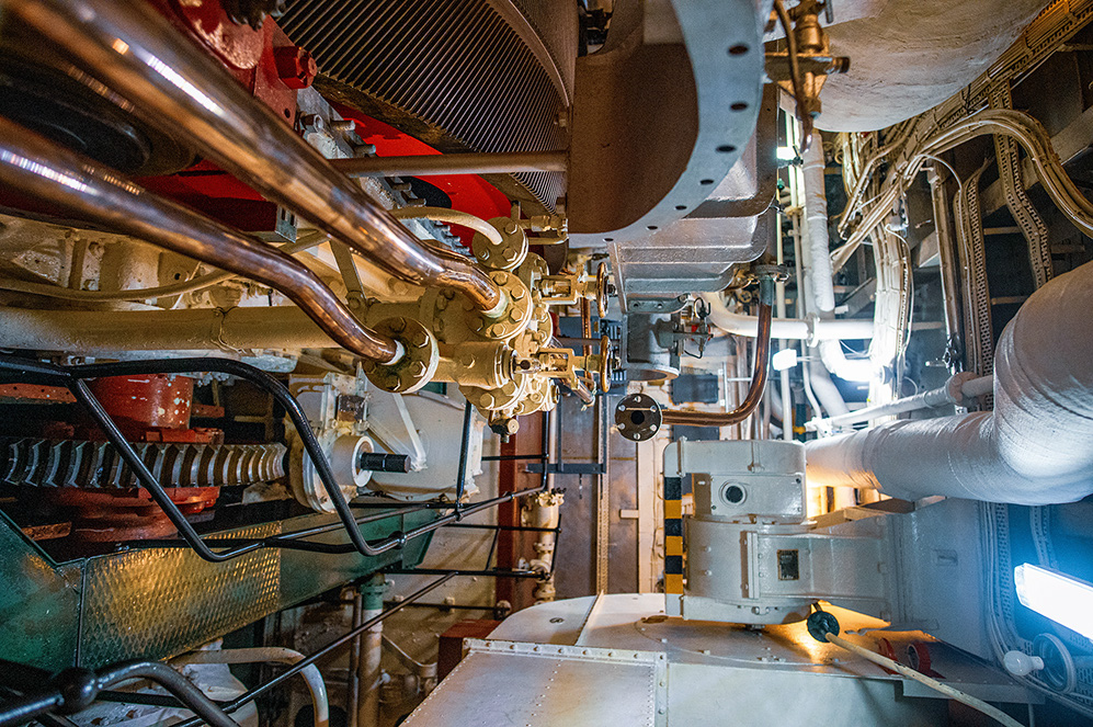 The image depicts the intricate inner workings of a complex mechanical system, likely the engine room of a ship or industrial machinery. It showcases a variety of pipes, valves, and mechanical components, highlighting the detailed and robust engineering involved. The environment appears well-lit, emphasizing the maintenance and operational readiness of the machinery.