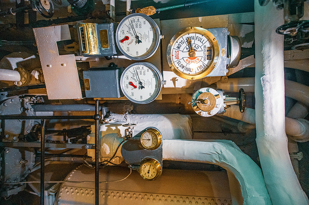 The image shows a complex industrial setup with multiple gauges and valves. There are three prominent pressure gauges with needles pointing to different pressure levels. A large valve with a circular handle is labeled with instructions such as 'FULL,' 'SLOW,' 'STOP,' and directional arrows. The setup includes various pipes and metal components, indicating a system likely used for monitoring and controlling pressure in an industrial environment.