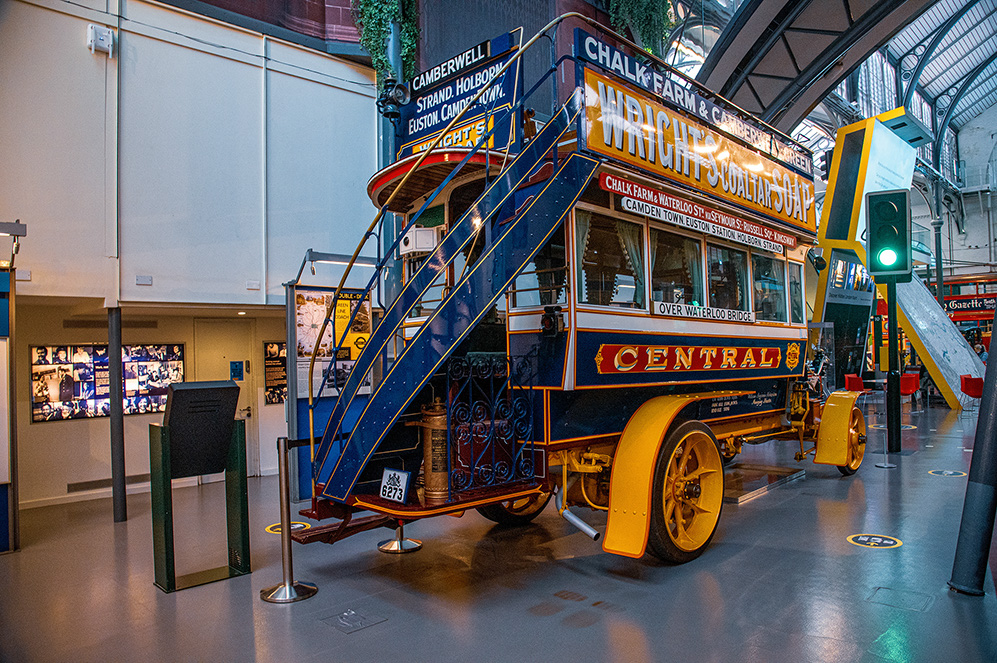 The image depicts a vintage double-decker tram on display in a museum. The tram, labeled 'CENTRAL,' is brightly colored with yellow and blue, and features a staircase for accessing the upper deck. The tram is surrounded by informational displays and barriers, indicating it is part of an exhibit. The setting appears to be indoors, with high ceilings and modern lighting. The tram's design and signage suggest it is a historical piece, likely from an era when such trams were used for public transportation.