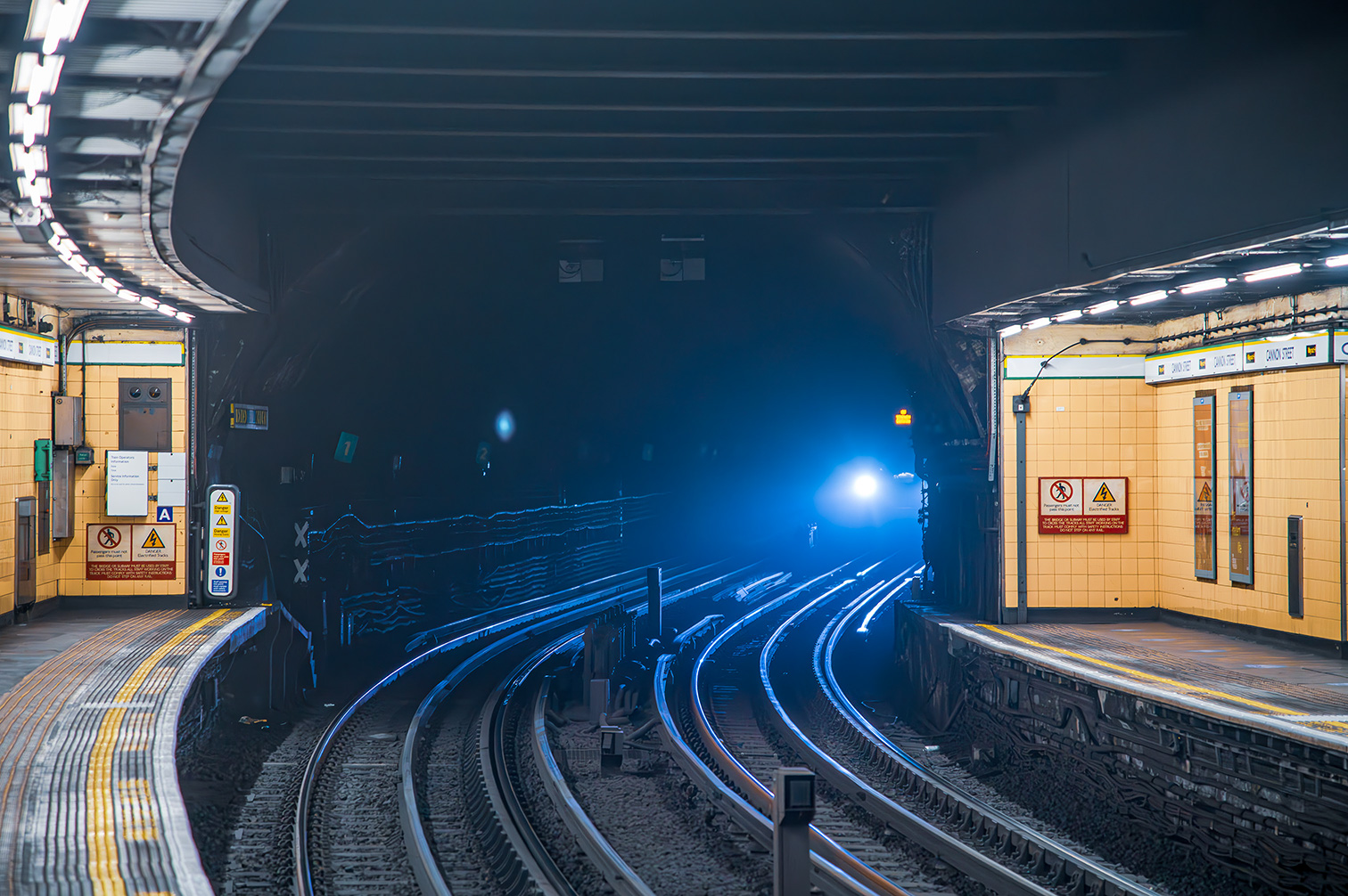 The image depicts an underground subway station with curved tracks extending into the distance. The station is well-lit with fluorescent lights on the ceiling, and there are various signs and notices on the walls. The tracks are bordered by yellow safety lines, and the station appears to be empty. The overall atmosphere is clean and orderly, with a sense of depth created by the converging tracks and the bright light at the end of the tunnel.