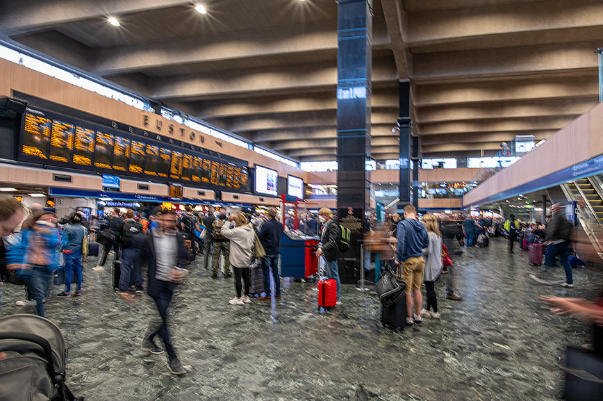 The image depicts a bustling scene at Euston railway station, with numerous people moving about, some carrying luggage. The station's departure board is prominently displayed, showing various train schedules. The architecture features a high ceiling with visible structural elements, and the area appears to be well-lit with natural light.