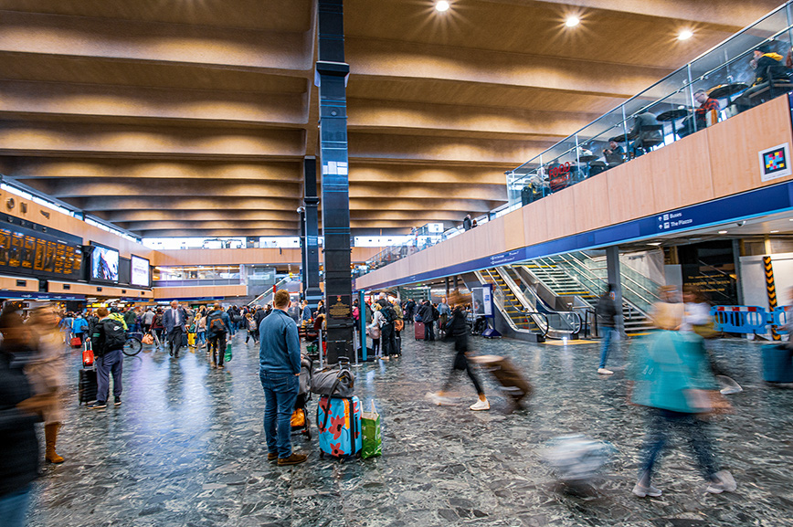 The image depicts a bustling airport terminal with numerous travelers moving about. The terminal features a high ceiling with wooden panels, large pillars, and a variety of shops and amenities on the upper level. There are escalators and stairs leading to the upper level, and a prominent display board showing flight information. The floor is made of polished stone, reflecting the overhead lights. The overall atmosphere is busy and dynamic, with people carrying luggage and moving in different directions