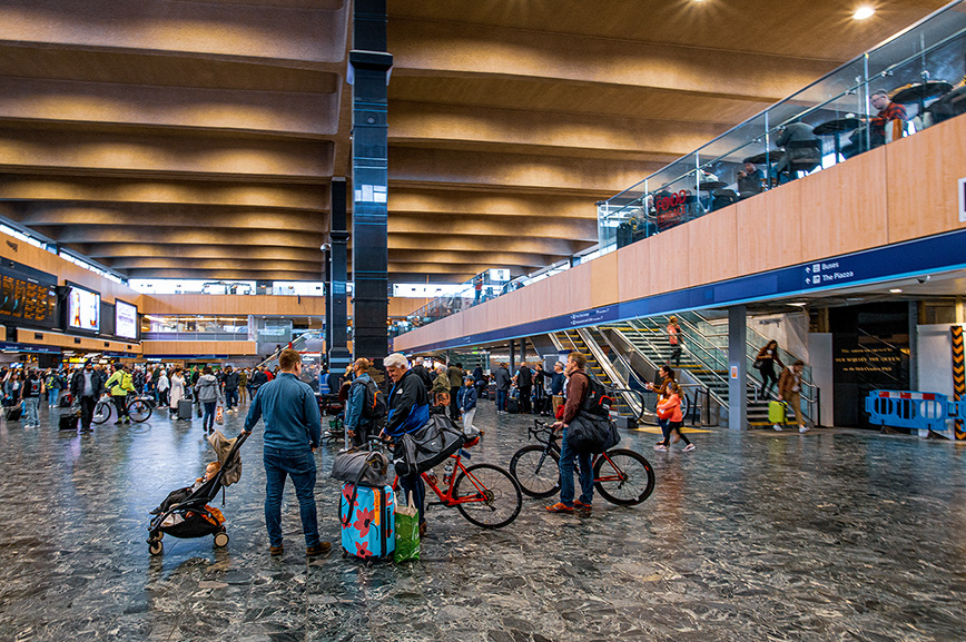 The image depicts a bustling indoor transportation hub, likely a train station, with people engaged in various activities such as standing, walking, and cycling. The architecture features a high ceiling with wooden panels and large columns. There are signs directing to different areas such as buses and platforms. The floor appears wet, suggesting recent cleaning or rain. Overall, it is a busy environment with a mix of travelers and commuters.