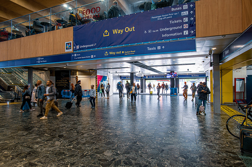 The image depicts a busy transportation hub, likely a train station or airport. There are clear signs directing passengers to various services such as buses, underground trains, taxis, and information desks. People are seen walking with luggage, indicating travel. The floor appears wet, suggesting recent cleaning or rain. The upper level has food and retail services.