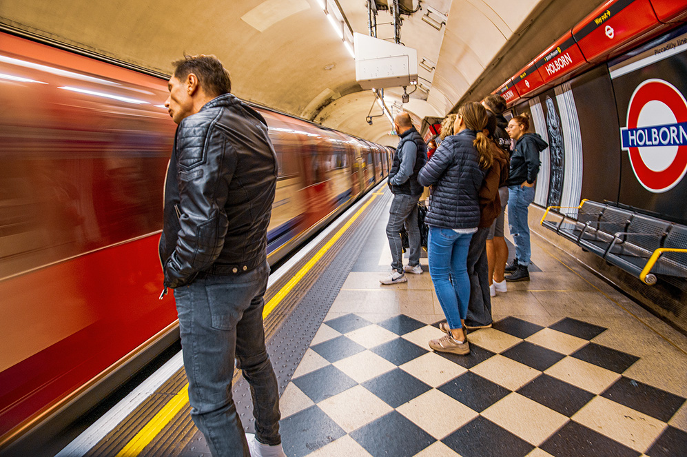 The image depicts a scene at the Holborn underground station. A train is moving quickly through the station, creating a blur on the left side of the image. Several people are waiting on the platform, some standing close to the edge and others near the benches. The station features a distinctive black and white checkered floor and a prominent Holborn station sign on the wall.