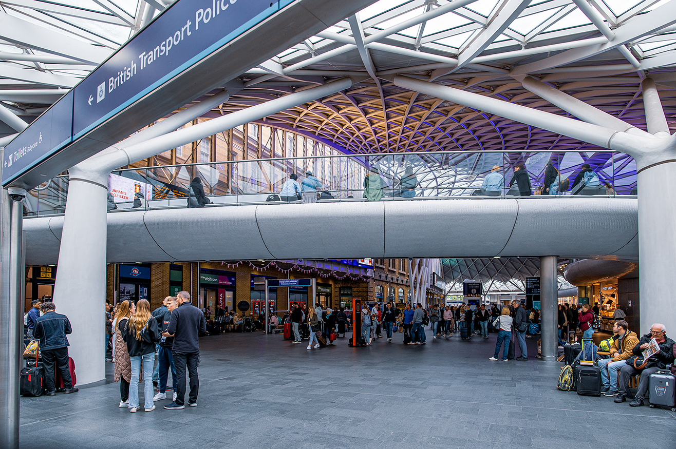 The image depicts a busy transportation hub, likely a train station, with a modern architectural design featuring a high ceiling with a complex network of metal beams and glass. There are numerous people, some standing and others walking, with luggage, indicating travel. Overhead signs point to the British Transport Police and other amenities. The station appears to be well-lit and bustling with activity.