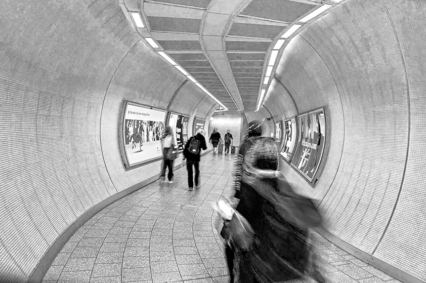 The image depicts a tunnel-like corridor with people walking through it. The corridor has a curved ceiling and walls lined with advertisements. The floor is tiled, and the scene is captured in black and white. The perspective is from someone walking through the corridor, with a slight motion blur effect. The people in the image appear to be in motion, heading towards the end of the corridor.