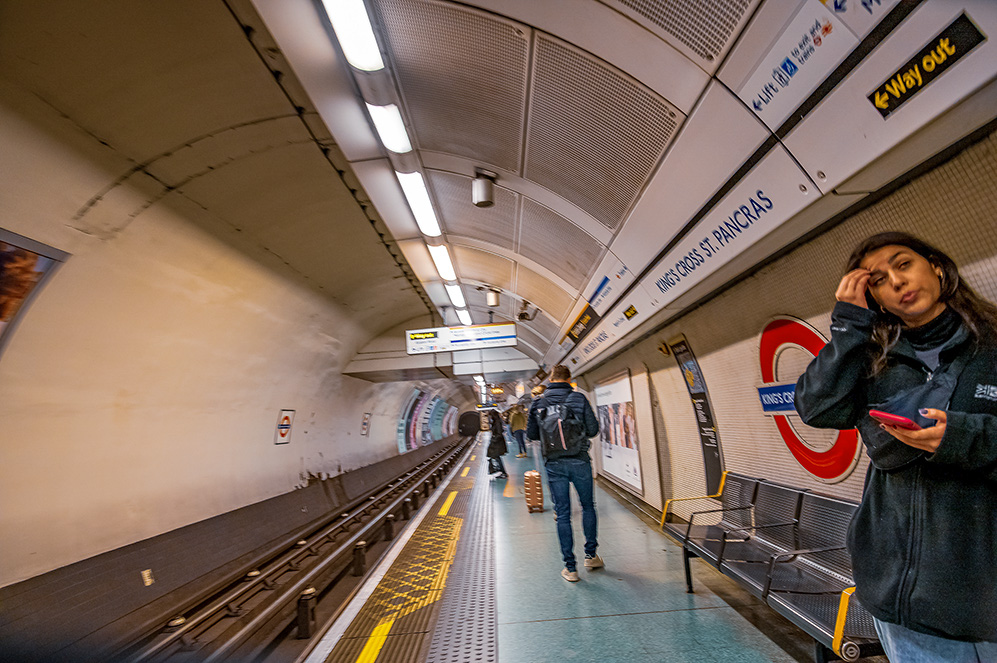 The image depicts a subway station platform, likely part of the London Underground system, as indicated by the iconic roundel sign. The station is Kings Cross St. Pancras, as seen from the signage. The platform is relatively empty, with a few people present. One person is walking away from the camera, while another person on the right is on the phone. The station is well-lit with fluorescent lights, and there are various advertisements and signs on the walls and ceiling.