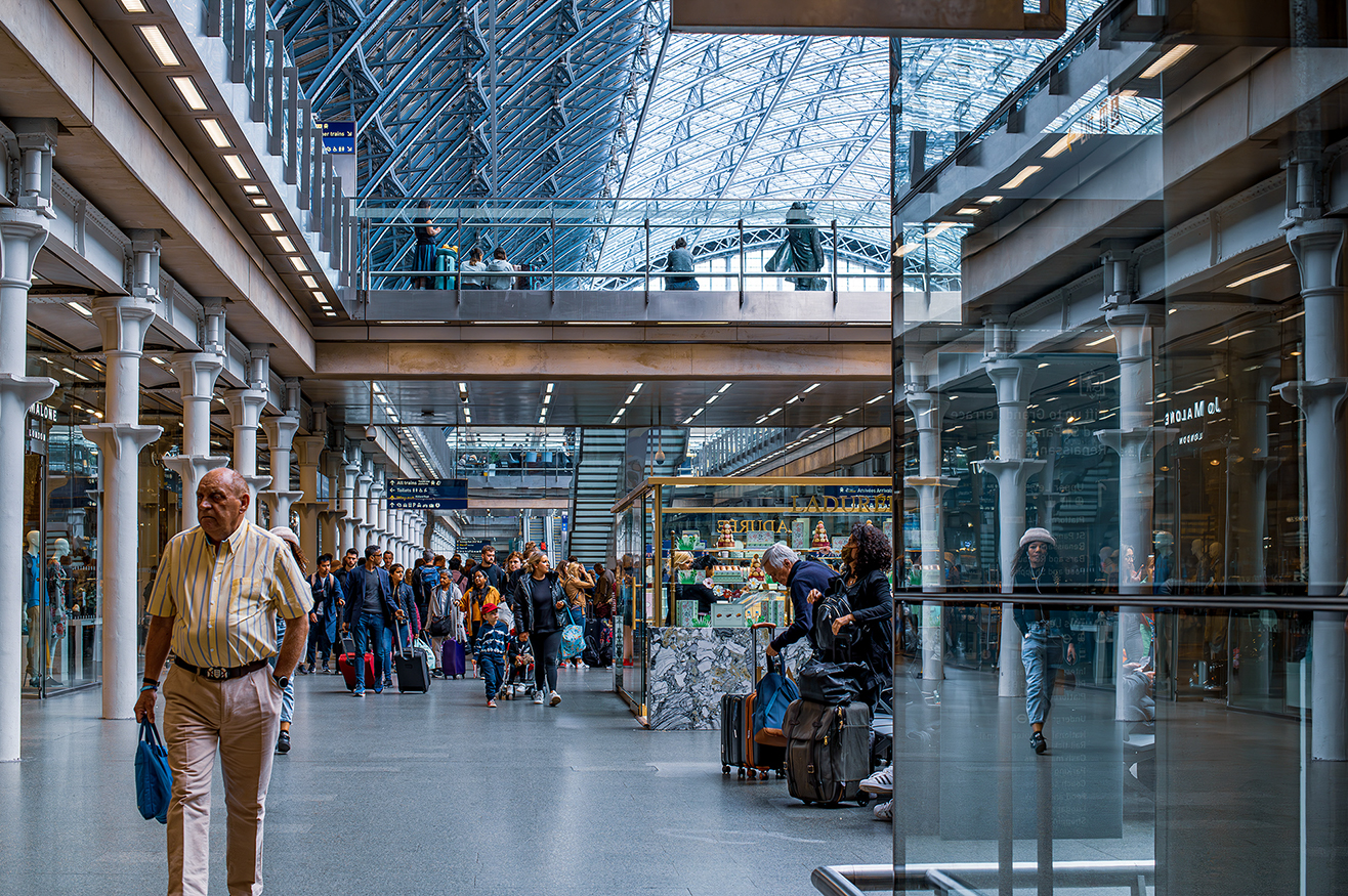 The image depicts a bustling scene at a modern train station. The station features a high, glass-paneled ceiling that allows natural light to flood the space, creating a bright and airy atmosphere. People are seen walking with luggage, indicating travel, and there are various shops and kiosks along the sides. The architecture includes sleek, contemporary design elements with glass and metal structures. Overall, it captures the dynamic and transient nature of a busy transportation hub.