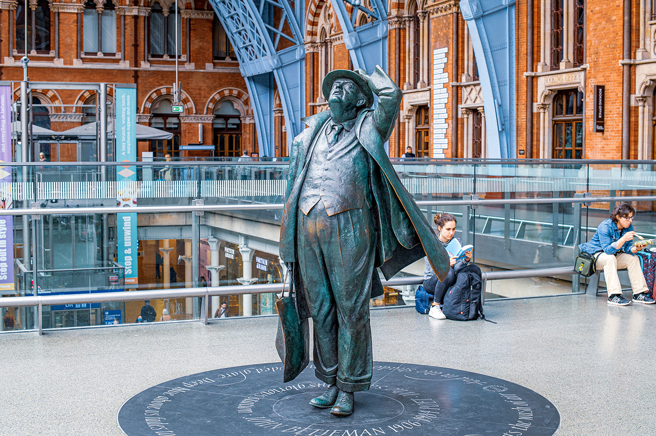 The image shows a statue of a man in a suit and hat, standing in a large, open indoor area with a high, arched ceiling and brick walls. The statue is positioned on a circular plaque with an inscription. The area around the statue is spacious, with a few people sitting and standing nearby. The architectural style of the building suggests it could be a train station or a similar public space.